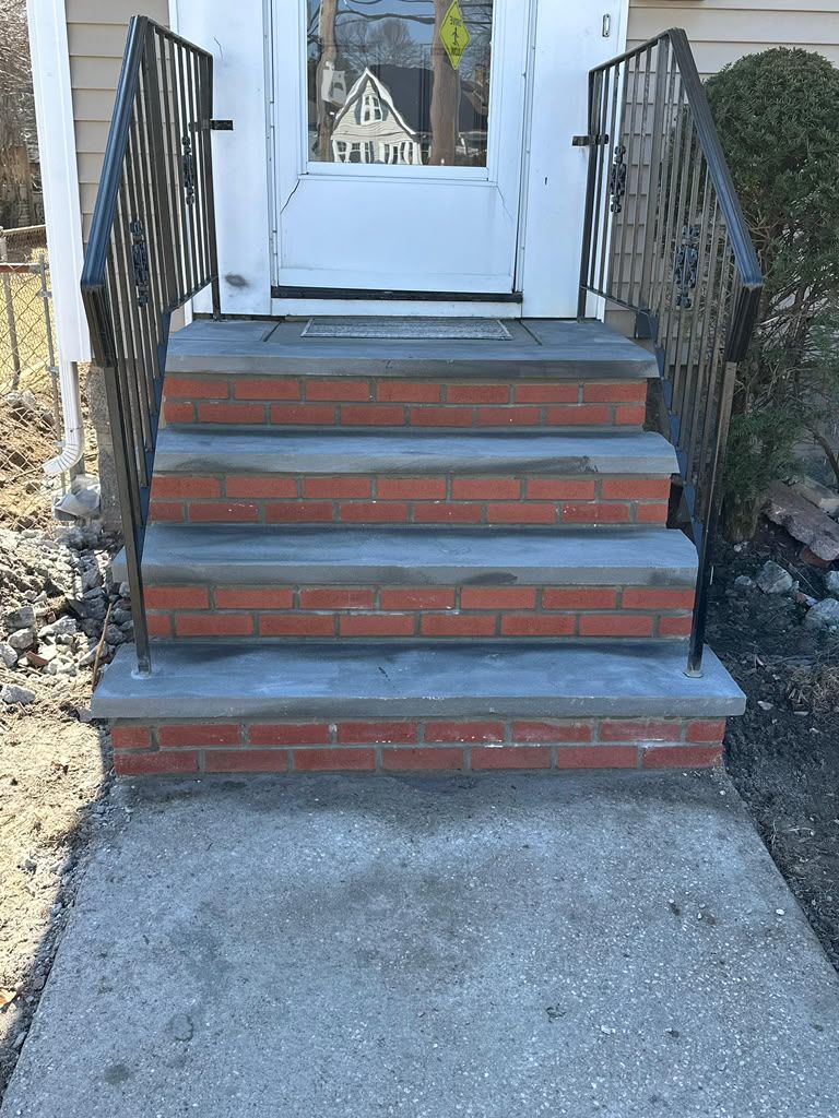 Front steps with black railings leading to a white door, with red brick risers and a concrete walkway.