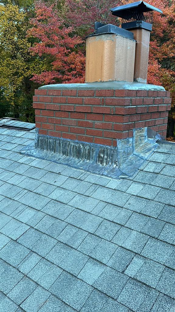 Brick chimney on a shingled roof with autumn trees in the background