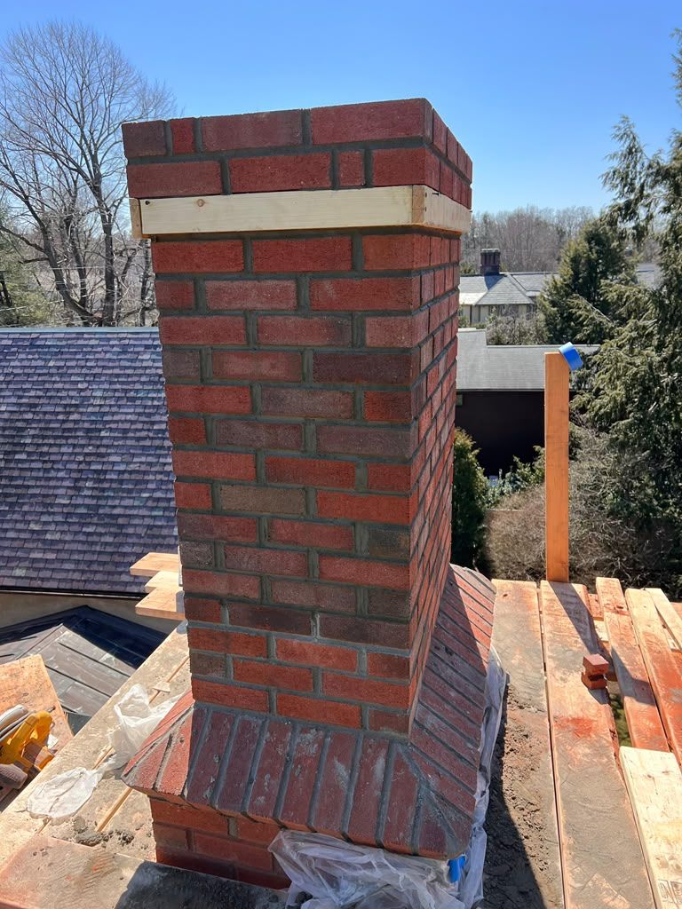 Red brick chimney under construction on a rooftop, with unfinished brickwork and nearby shingles.