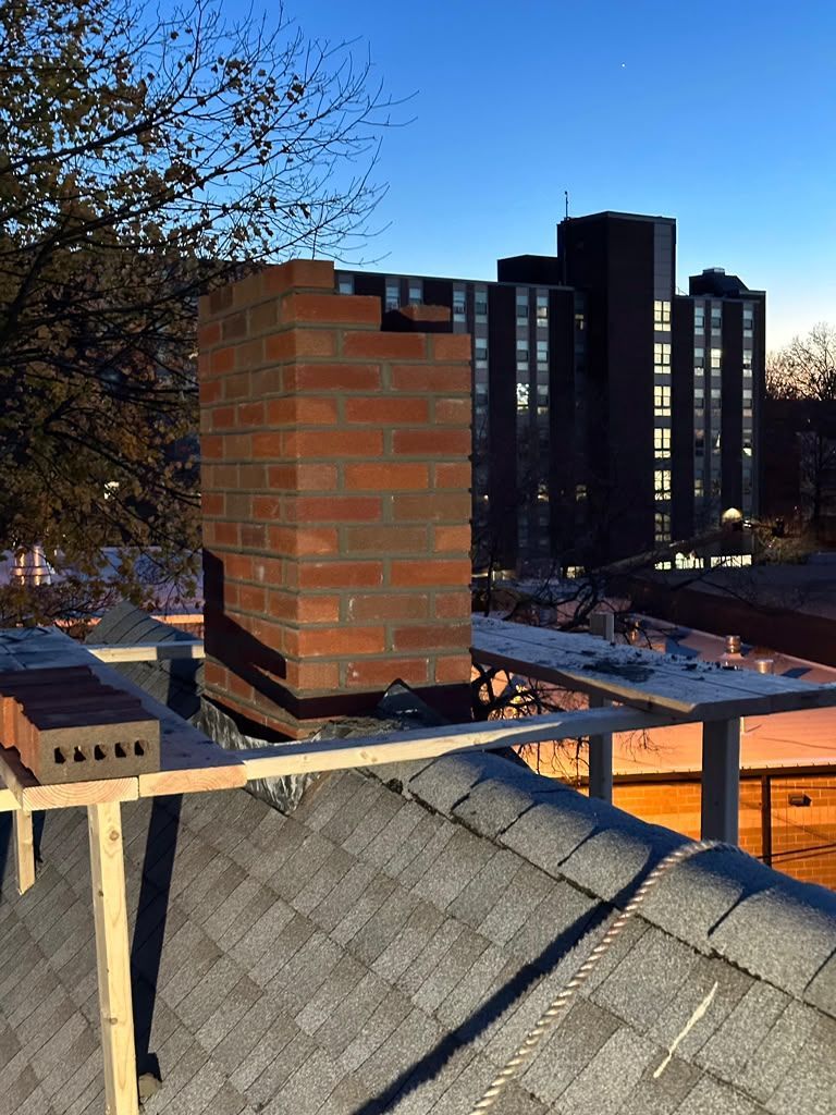 Brick chimney on a rooftop with shingles, overlooking tall apartment buildings at dusk.