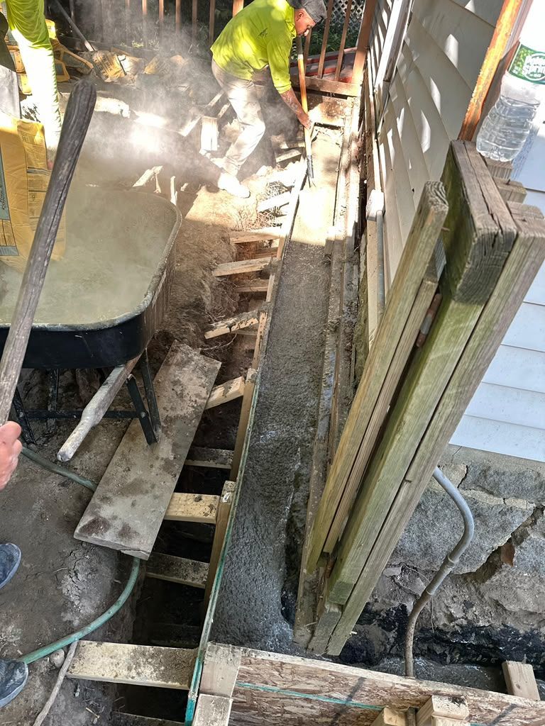 Worker repairing a narrow wooden trench beside a house, with a wheelbarrow and tools nearby.