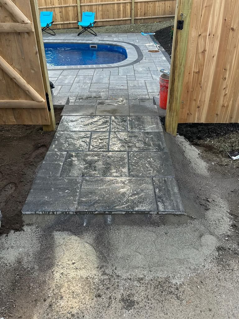 Stone patio path leading to a backyard pool, with wet pavers and a wooden fence gate.