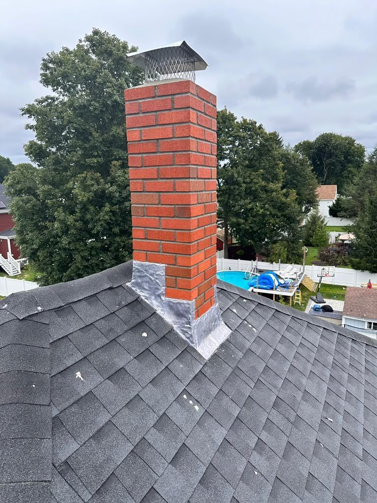 Brick chimney on a shingled roof with metal cap, surrounded by trees and cloudy sky.