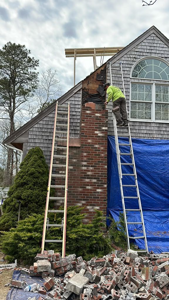 Workers repairing a brick house roof with ladders, blue tarp, and scattered debris below