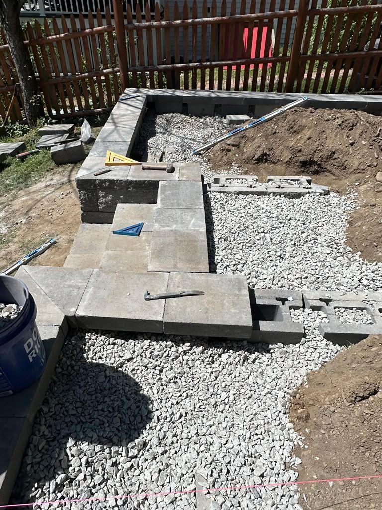 Backyard patio under construction with pavers, gravel, and a shovel near a wooden fence
