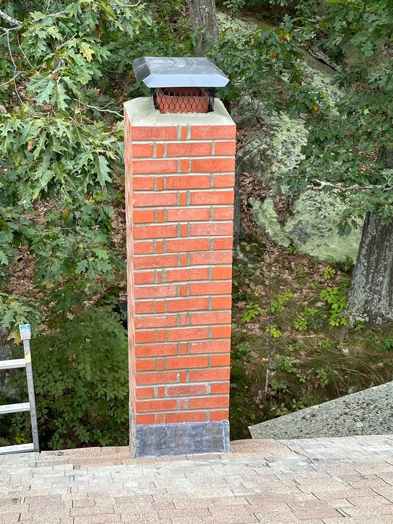Red brick chimney with metal cap on a rooftop, framed by leafy trees.