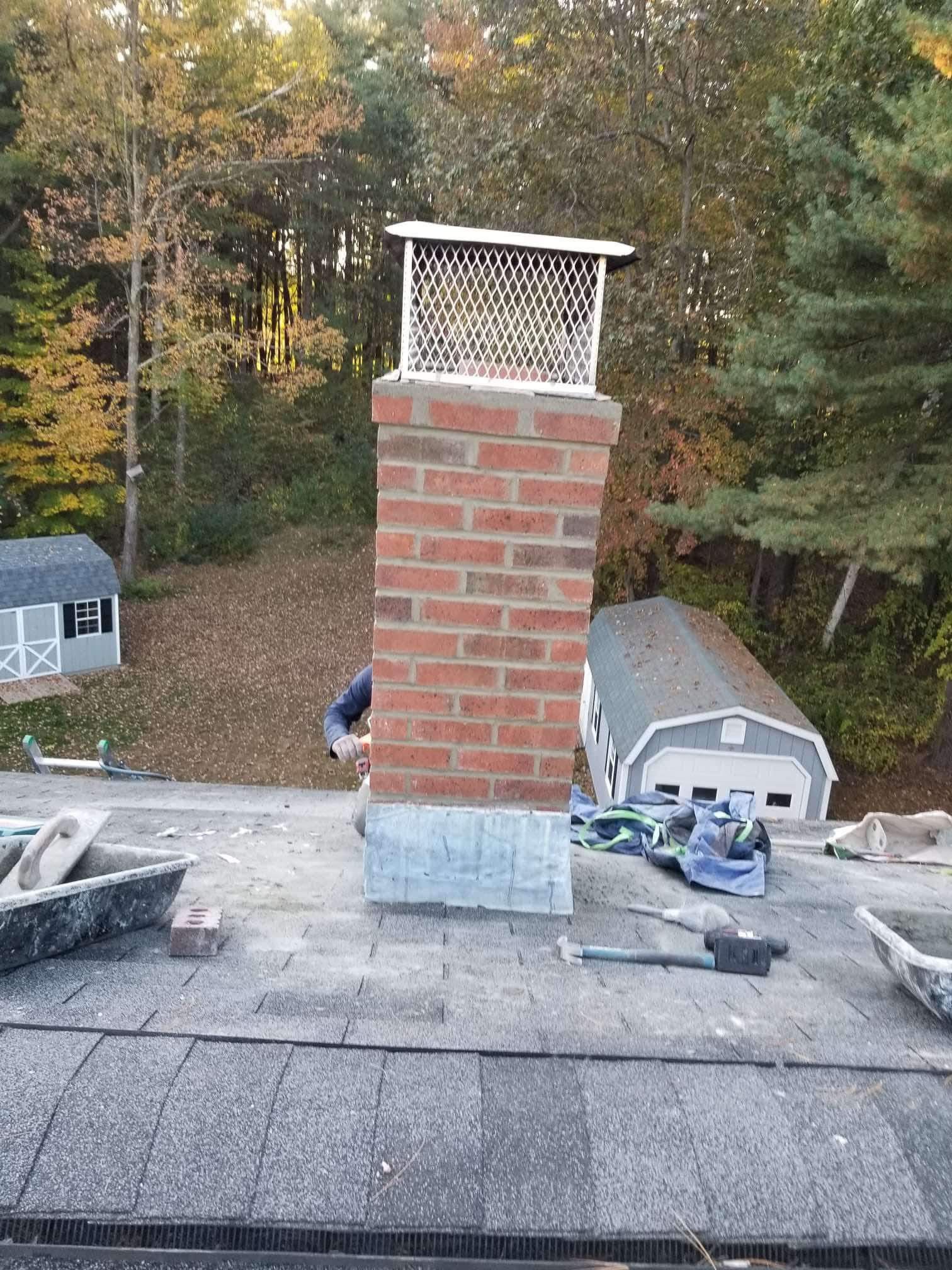 Brick chimney on a shingled roof with a metal cap, surrounded by trees and rooftop tools.