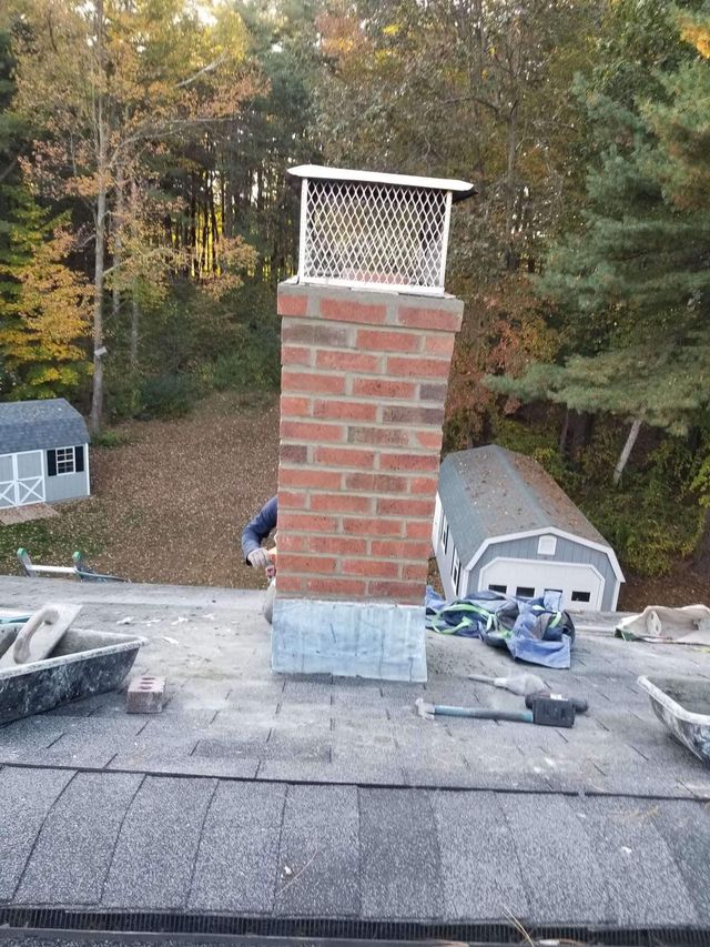 Brick chimney on a shingled roof with a metal cap, surrounded by trees in the background.