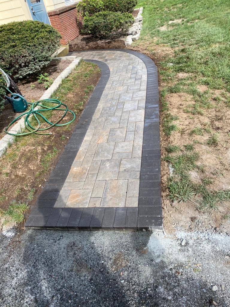 Curved brick garden walkway leading to a patio, bordered by grass and shrubs, with a garden hose on the left.