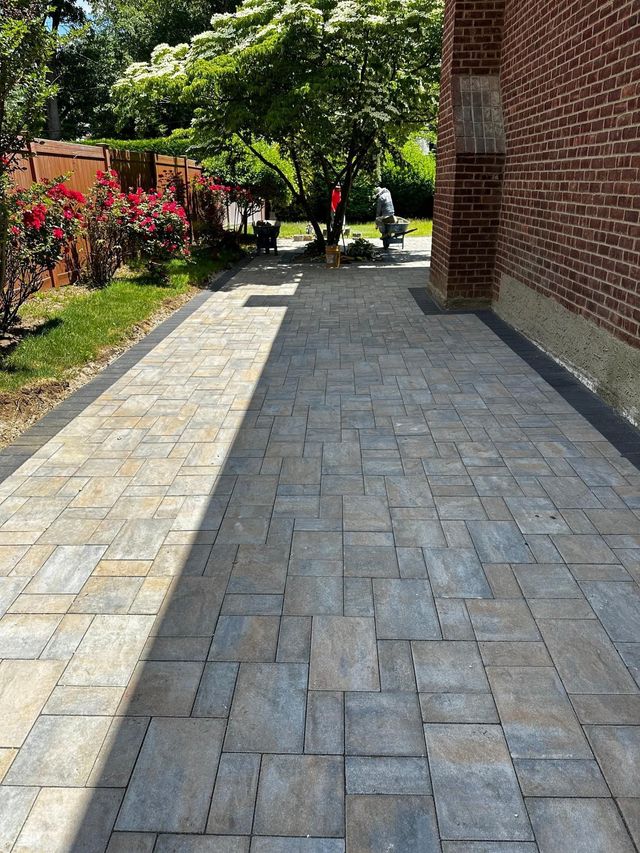 Brick walkway beside a garden and building, leading toward a shaded tree-lined path.