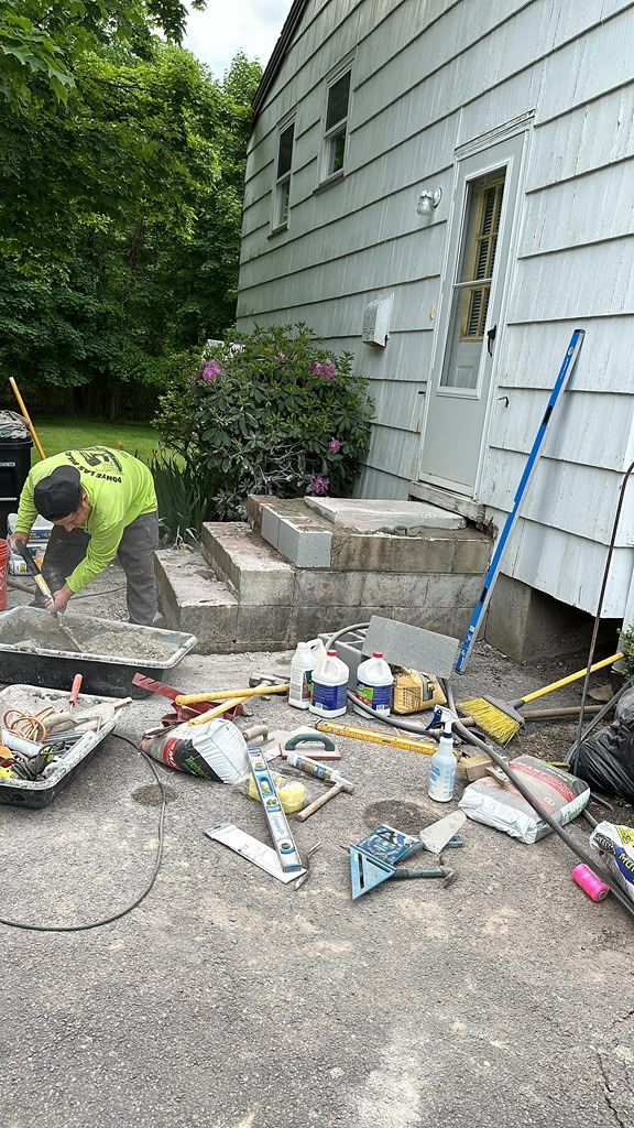 Worker in neon green shirt shovels debris beside a house with scattered construction materials.