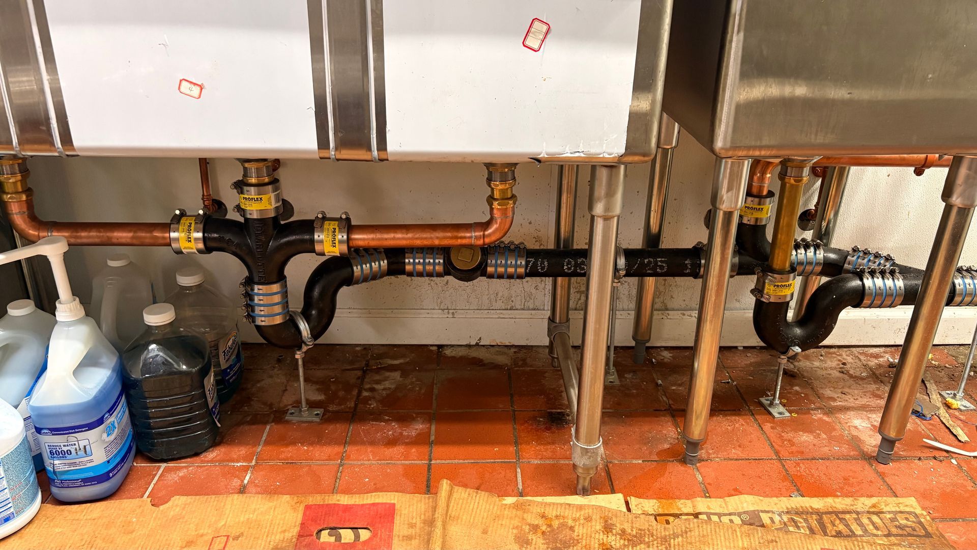 Plumbing under a triple sink. Copper pipes, black fittings, and bottles on red tile floor.