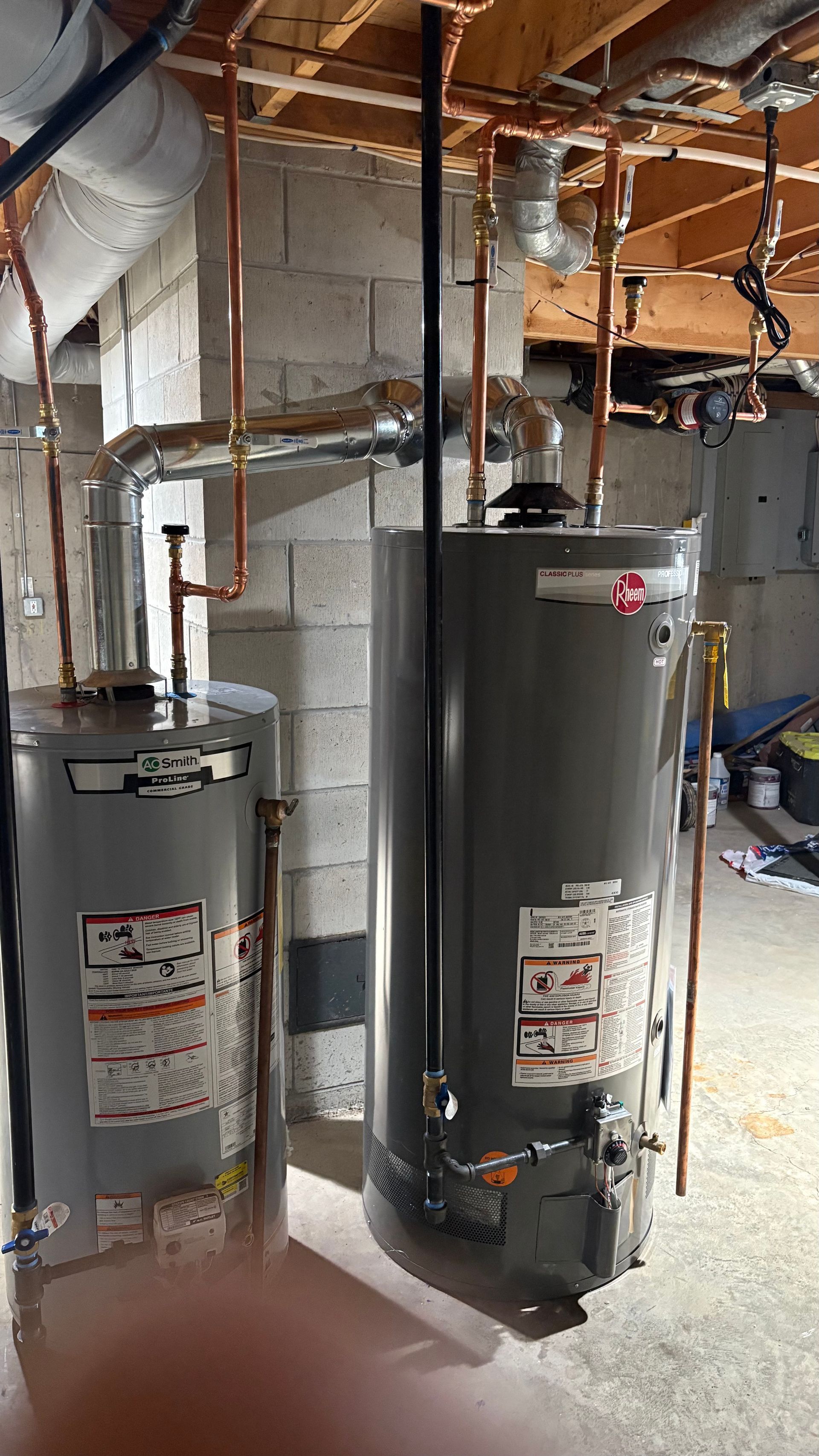 Two gray water heaters in a basement. Copper pipes and black flue are visible.