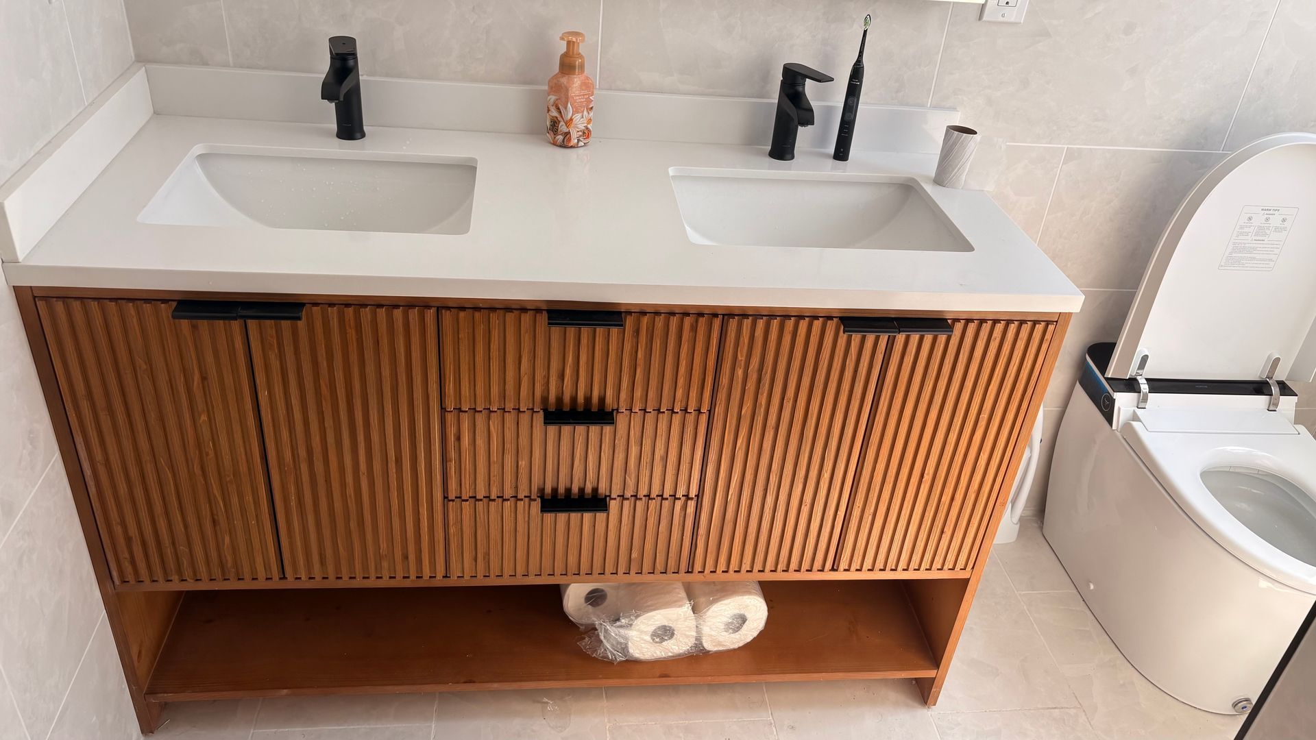 Wooden bathroom vanity with double sinks, black faucets, and an open lower shelf. Toilet on the right.