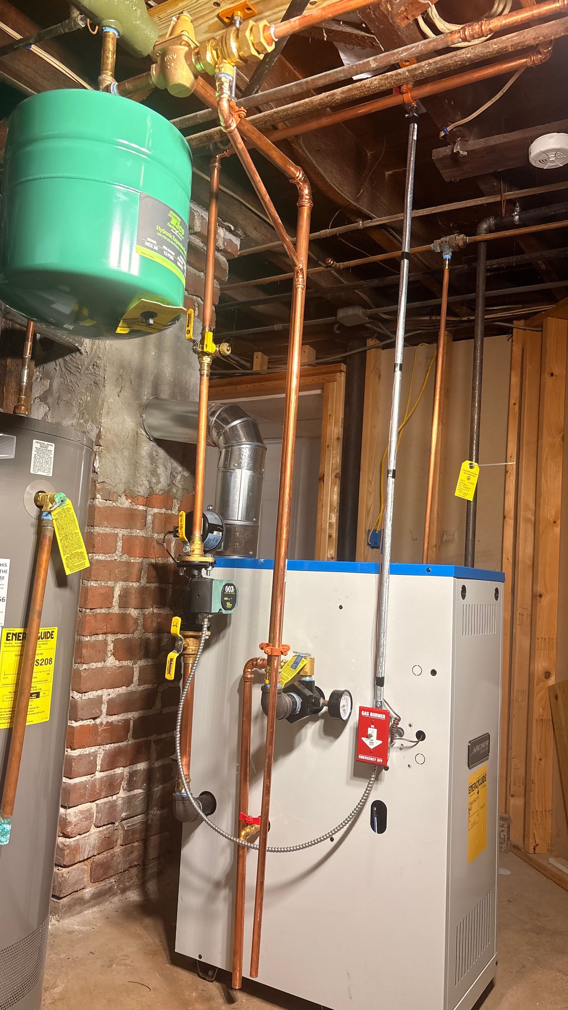 A boiler system with copper pipes, a green expansion tank, and a red fire alarm in a utility room.