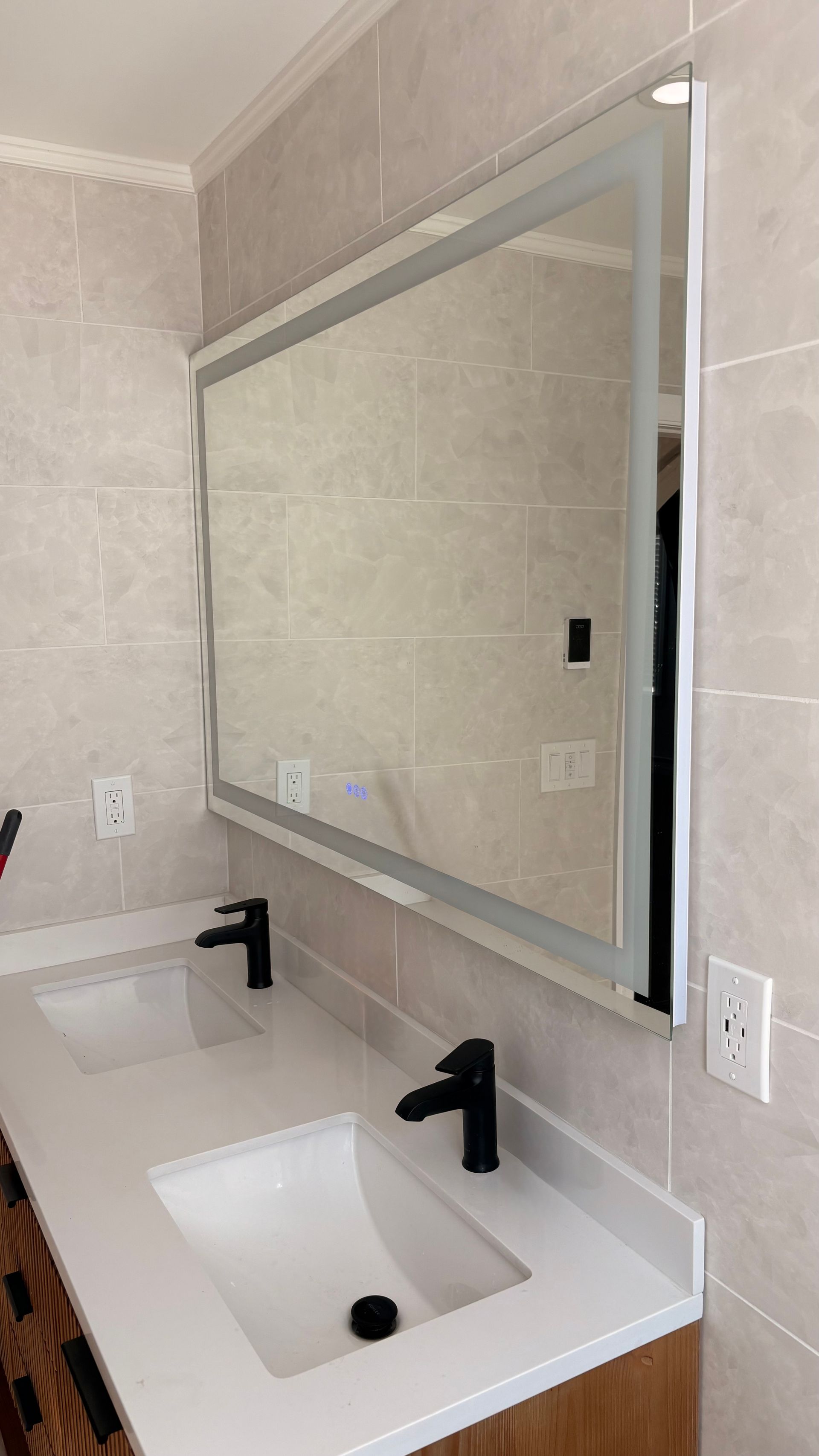 Bathroom with double sinks, large illuminated mirror, black faucets, and light-colored tile walls.