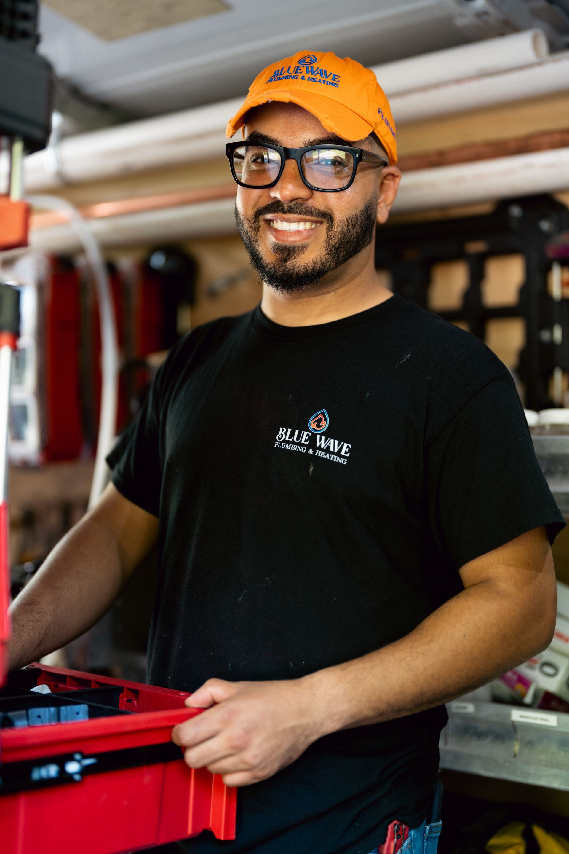 Man in black t-shirt and orange cap, wearing glasses, smiling, holding red toolbox in workshop.