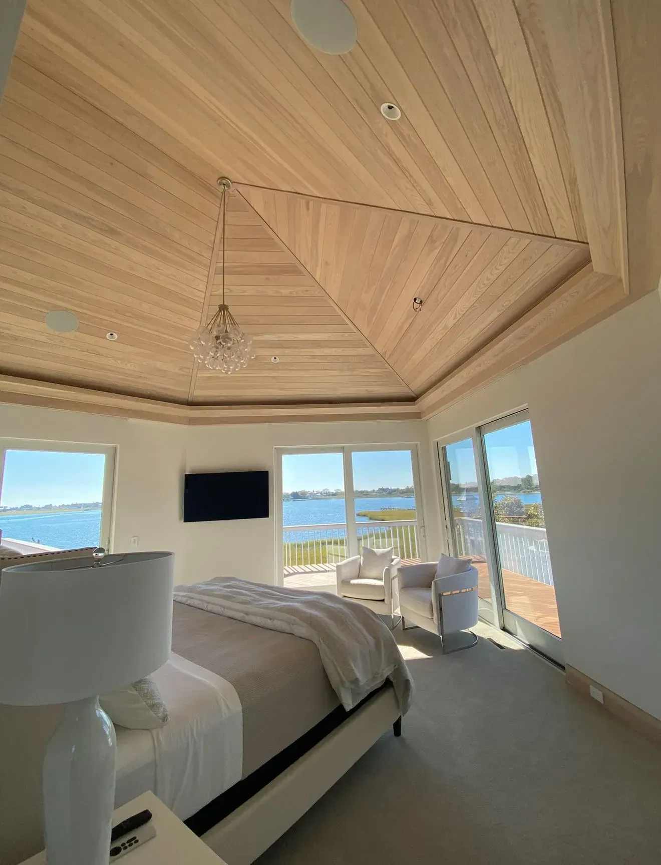 Bedroom with wood-paneled ceiling, large windows overlooking water, bed, and chairs.