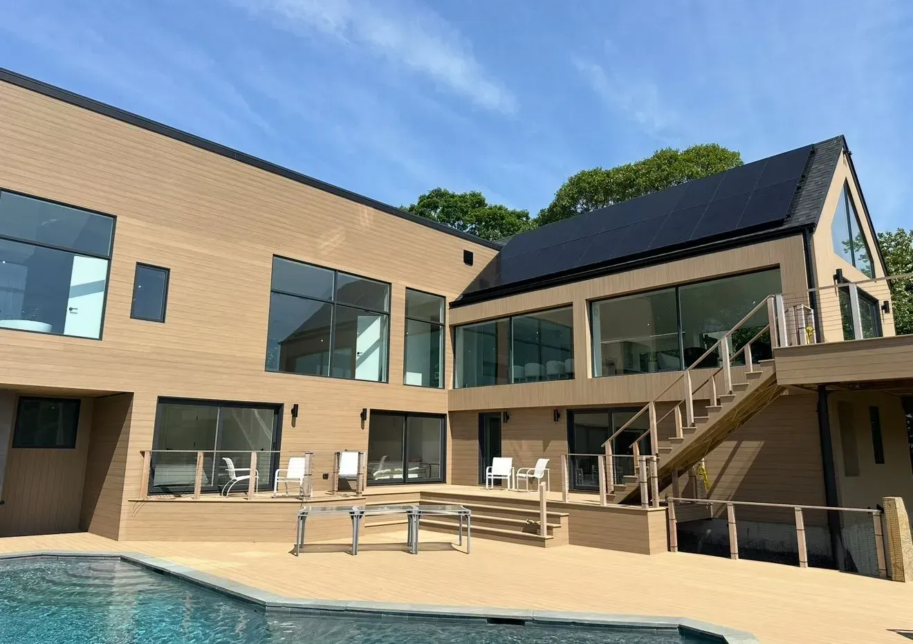 Modern beige house with large windows, pool, and wooden deck under a blue sky.