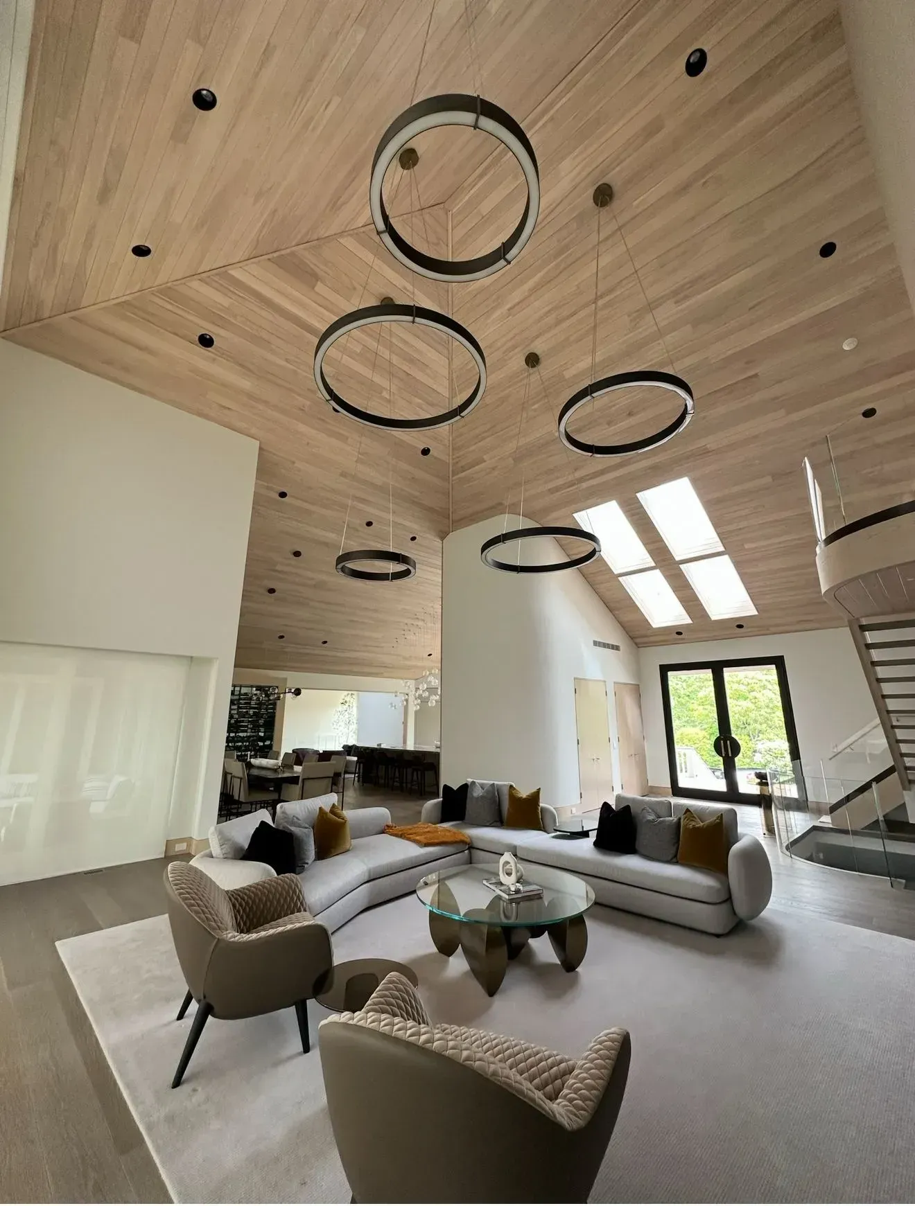 Modern living room with light wood ceiling, circular lights, sofas, chairs, and natural light.