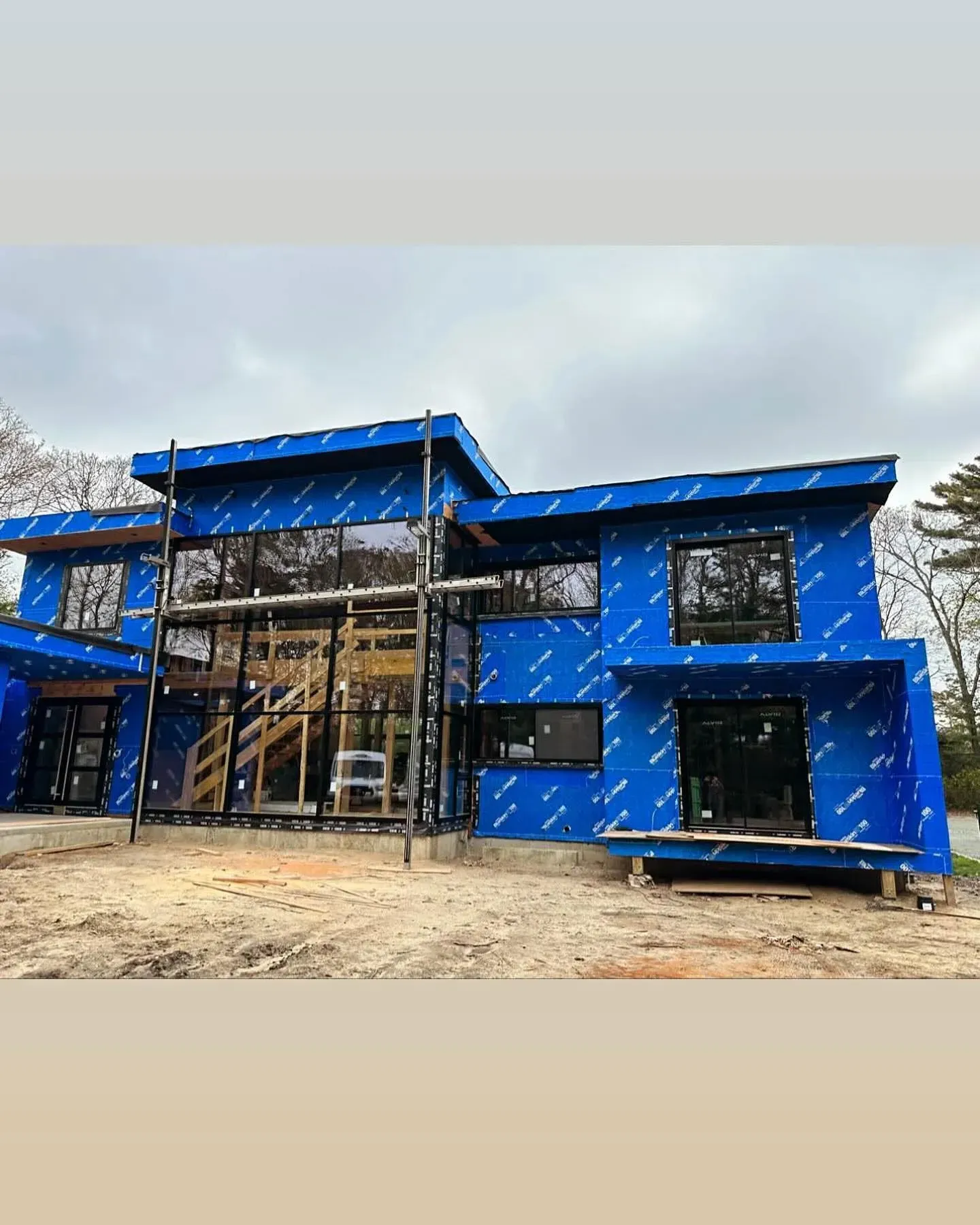 Modern house under construction; blue protective wrap covers exterior walls; large windows and flat roof.
