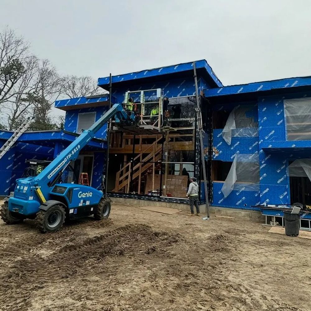 Construction site with a blue Genie lift and workers on a partially built modern house, covered in blue sheathing.