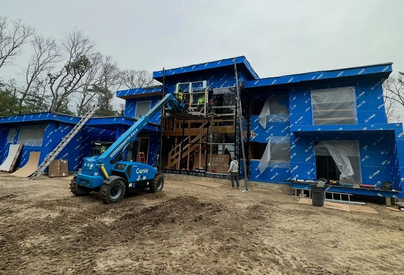 Construction site with a blue lift and workers installing windows in a modern home wrapped in blue protective sheathing.