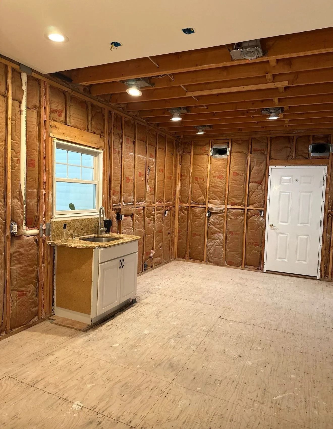 Interior of a room under construction; insulation visible between wood studs, sink, white door, and recessed lighting.