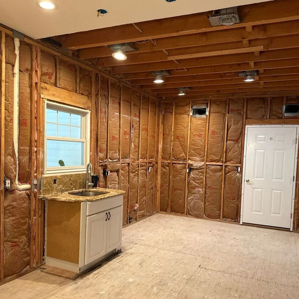 Interior room under construction, with exposed wood framing, insulation, a window, sink, and door.