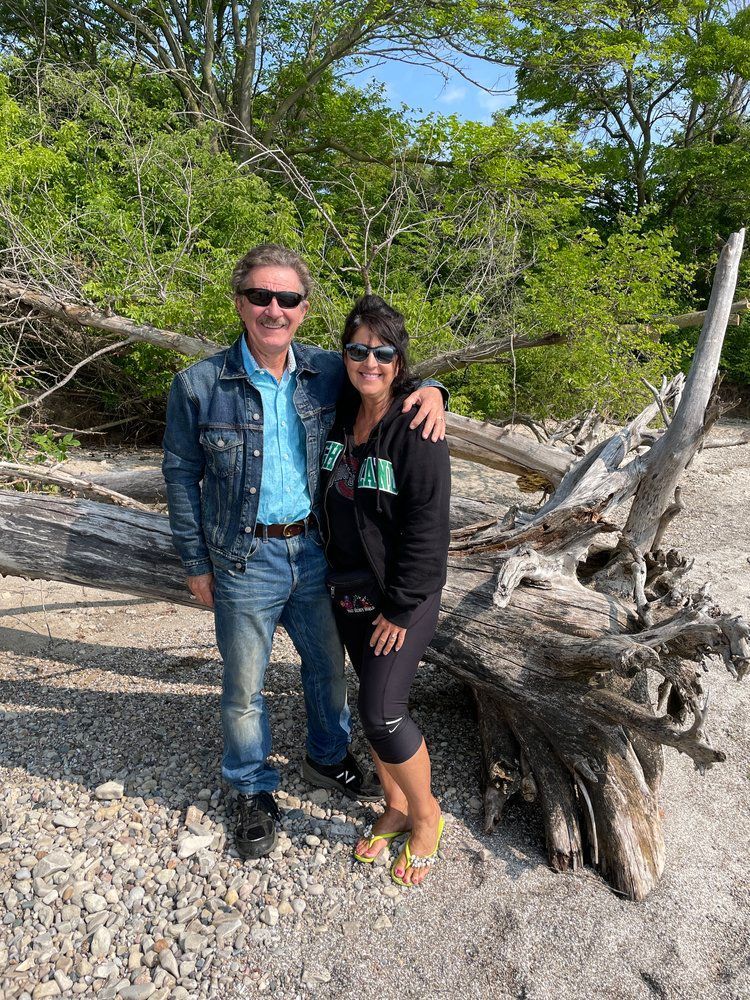 Two people standing on a rocky beach by driftwood and trees, smiling in bright sunlight