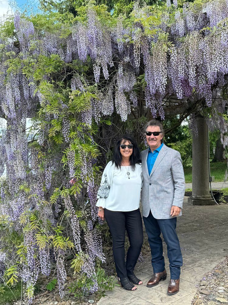 Two people standing under hanging purple wisteria in an outdoor garden, both wearing sunglasses and smiling.