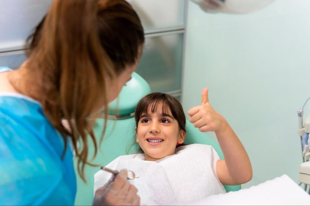 Child in a dental chair giving a thumbs-up while a dentist works nearby