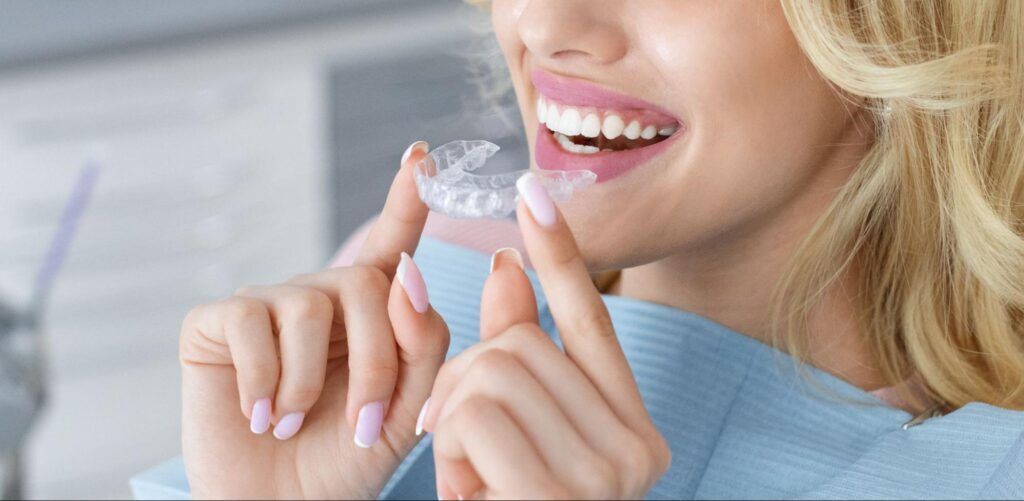 Person holding a clear aligner near their smiling mouth in a bright clinical setting
