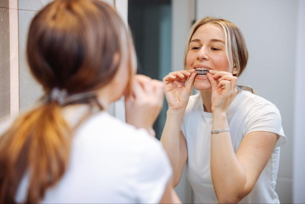 Woman flossing teeth in a bathroom mirror, wearing a white shirt.