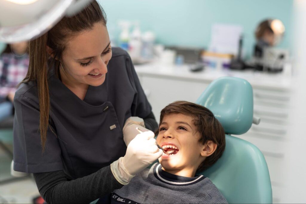 Dentist examining a smiling child in a dental chair with a gloved hand and mirror