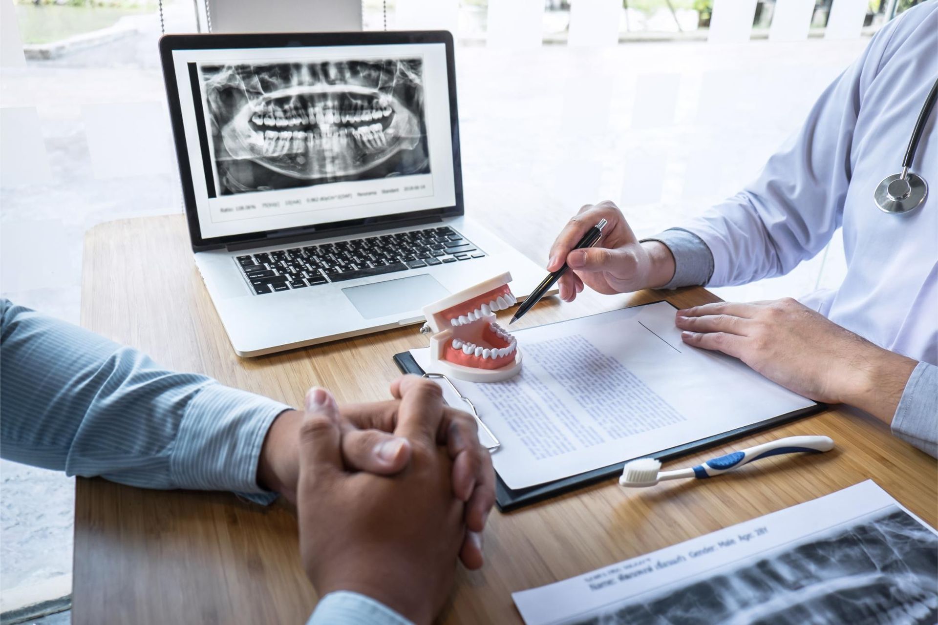 Doctor showing dental model to patient at desk with X-ray on laptop