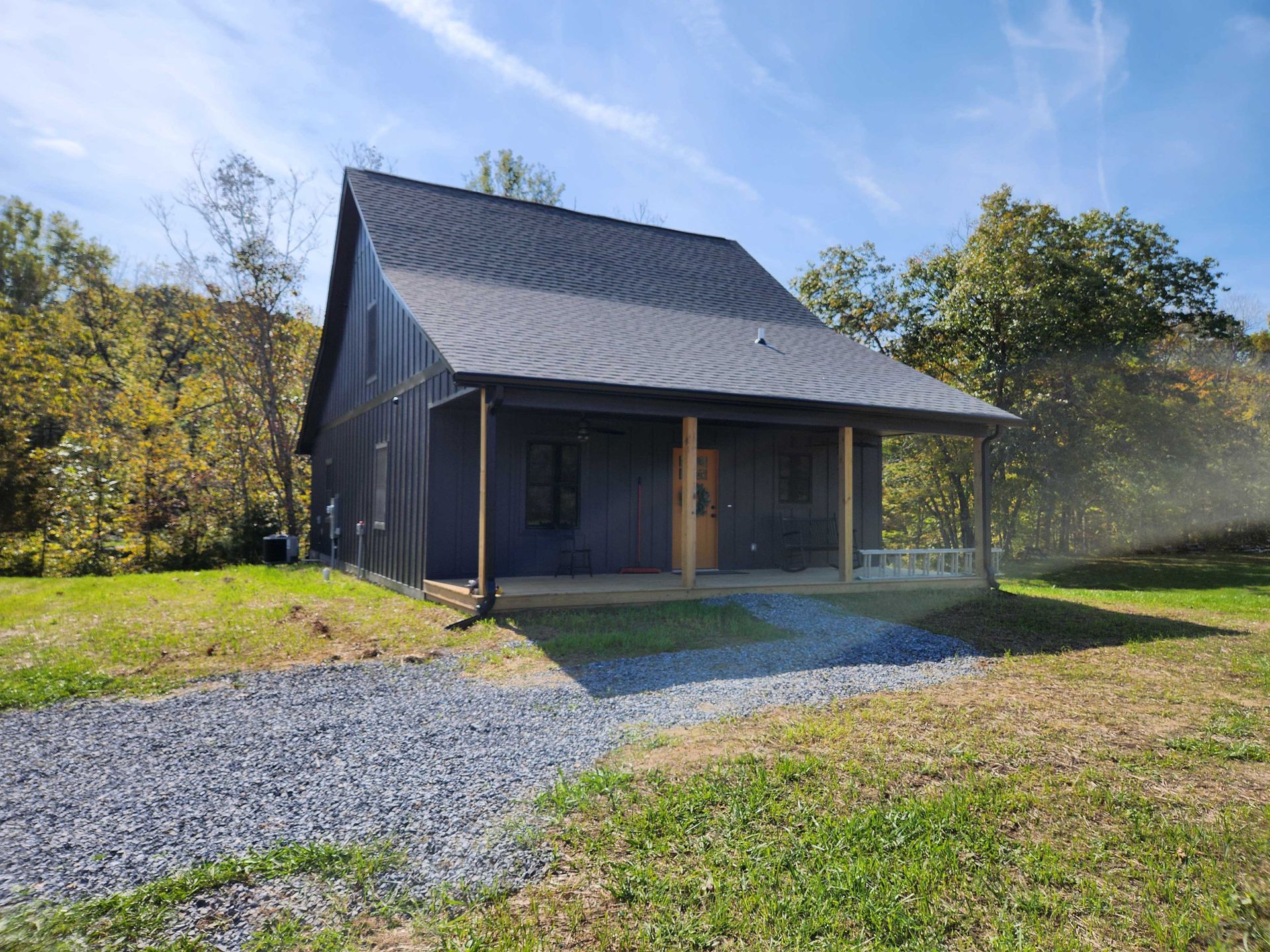 Small gray cabin with porch and gravel driveway in a grassy setting with trees.