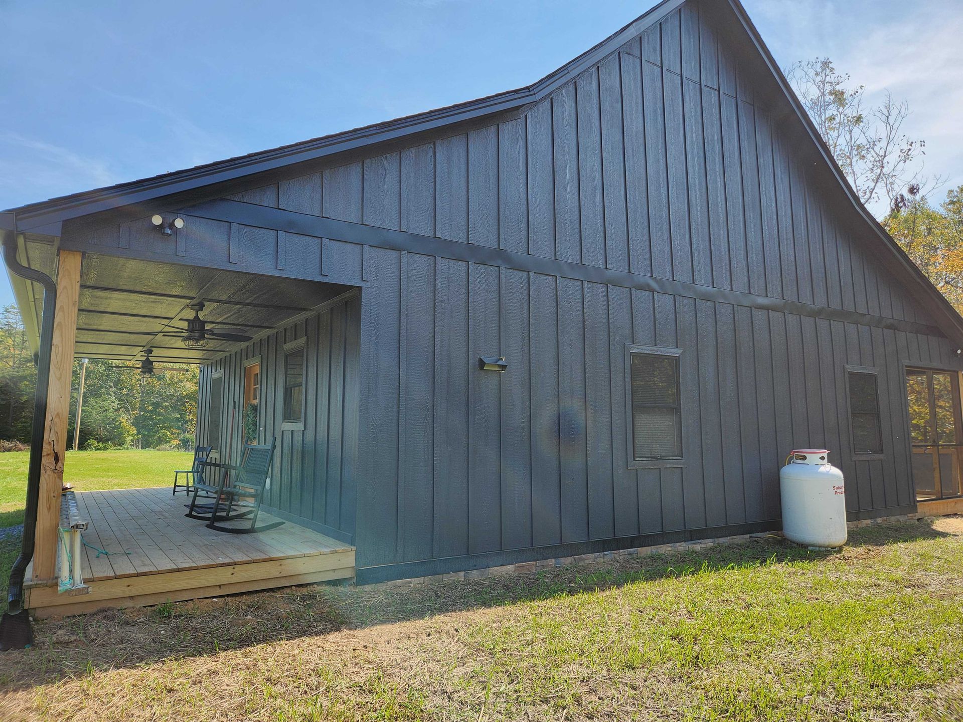 Black-sided cabin with a porch, green lawn, and propane tank on a sunny day.