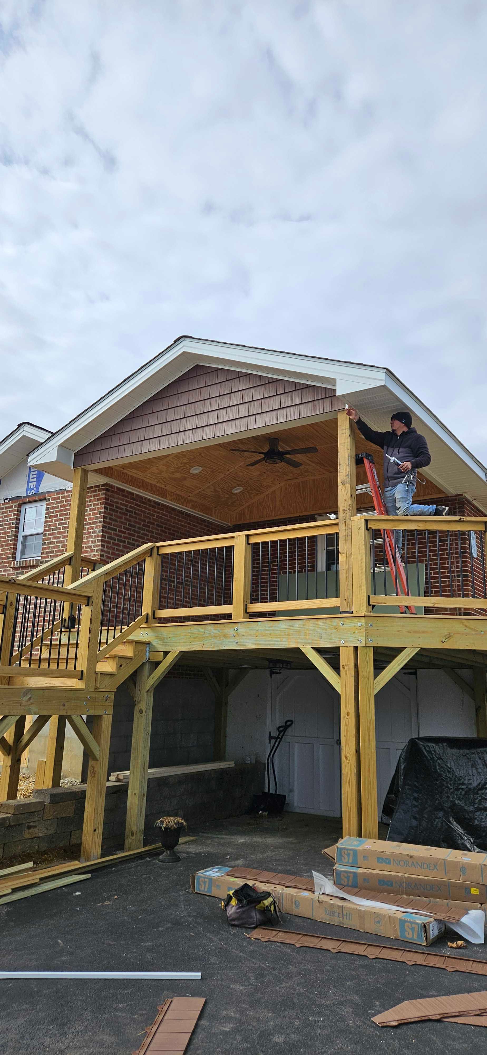 Wooden deck under construction next to a house with a person working on the roof. Cloudy sky.