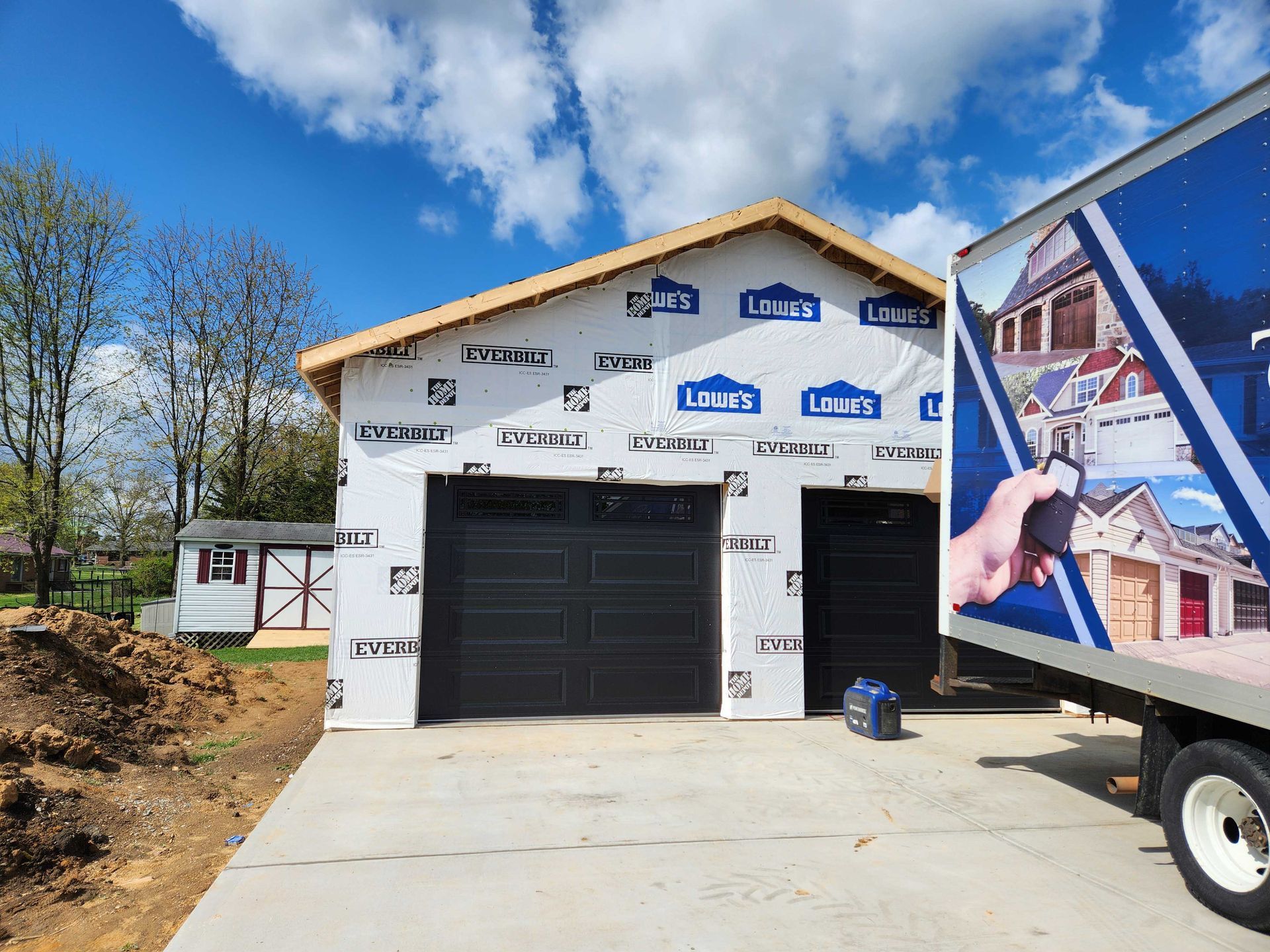 New garage under construction with two black garage doors and a Lowe's tarp. A box truck is parked next to it.