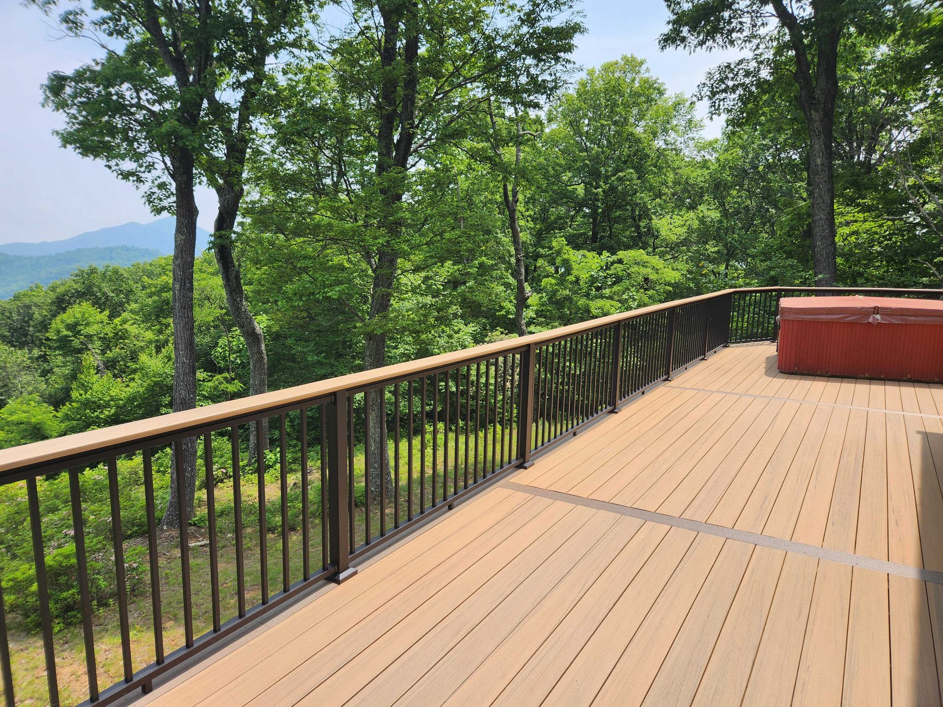 Deck with brown composite decking, black railing, and red hot tub, surrounded by trees with a mountain view.