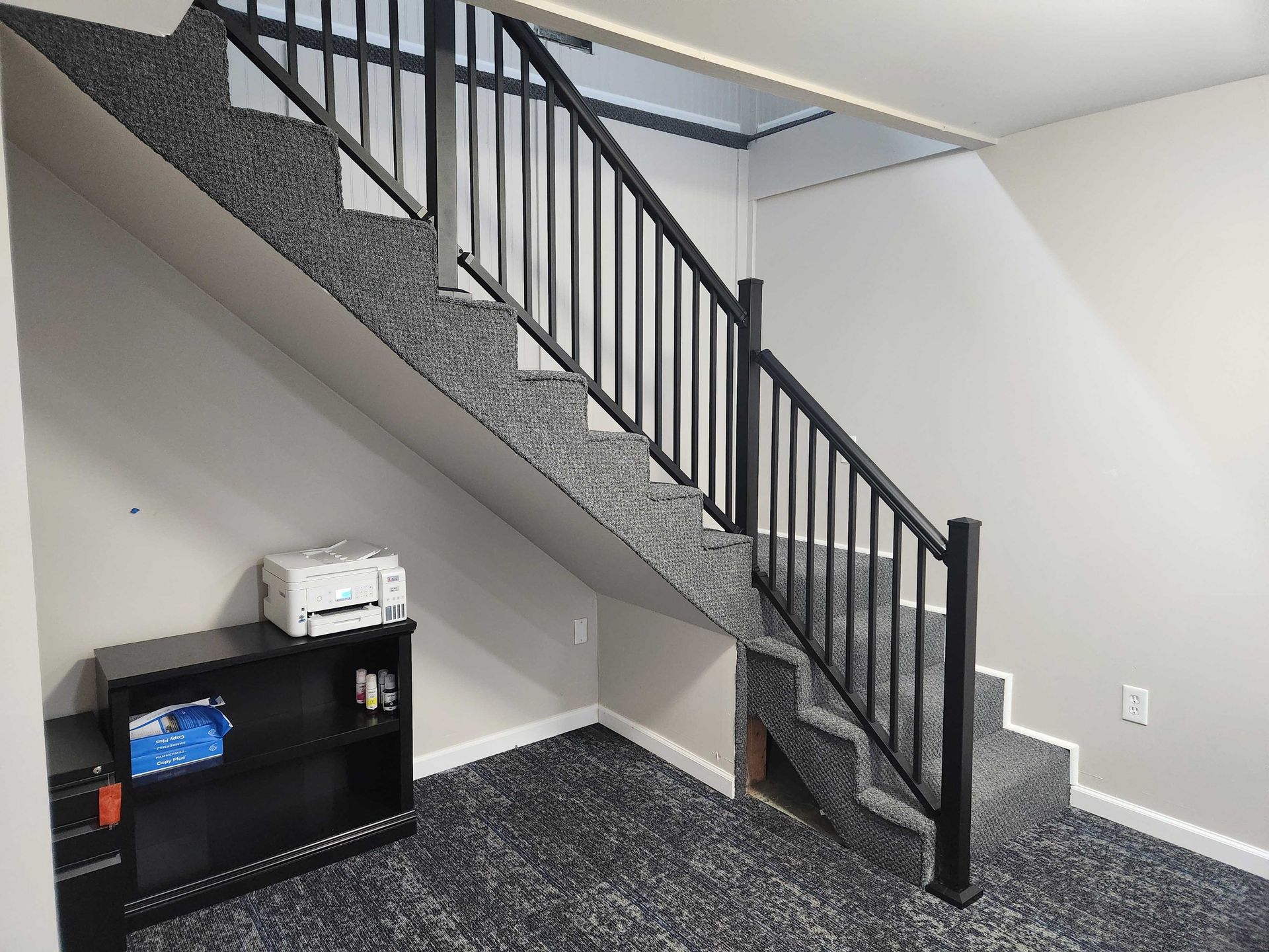 Staircase with black railing and carpeted steps. A small bookshelf and printer are visible below the stairs.