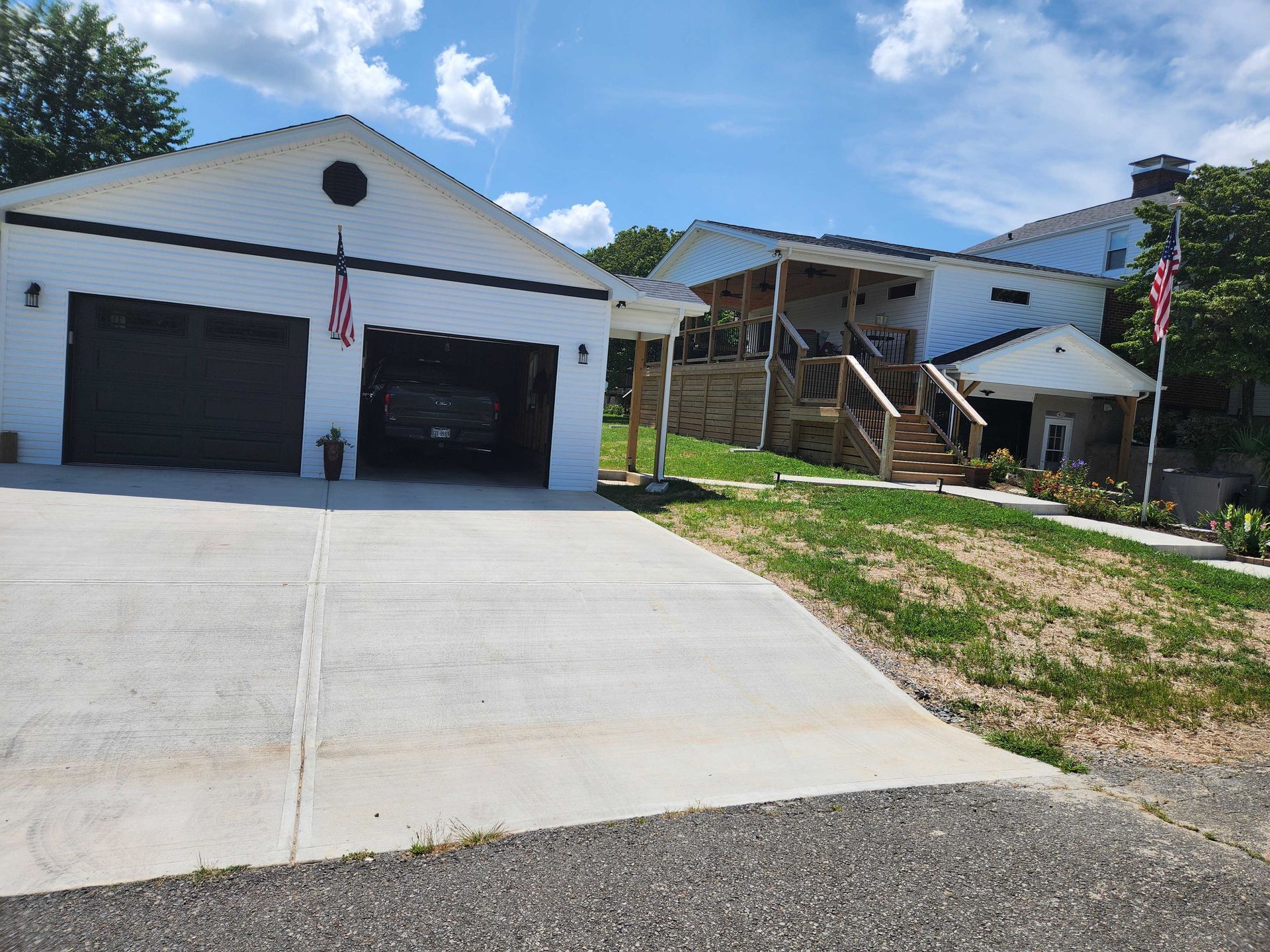 White house with black garage doors, car inside. American flags, concrete driveway, and grassy yard.