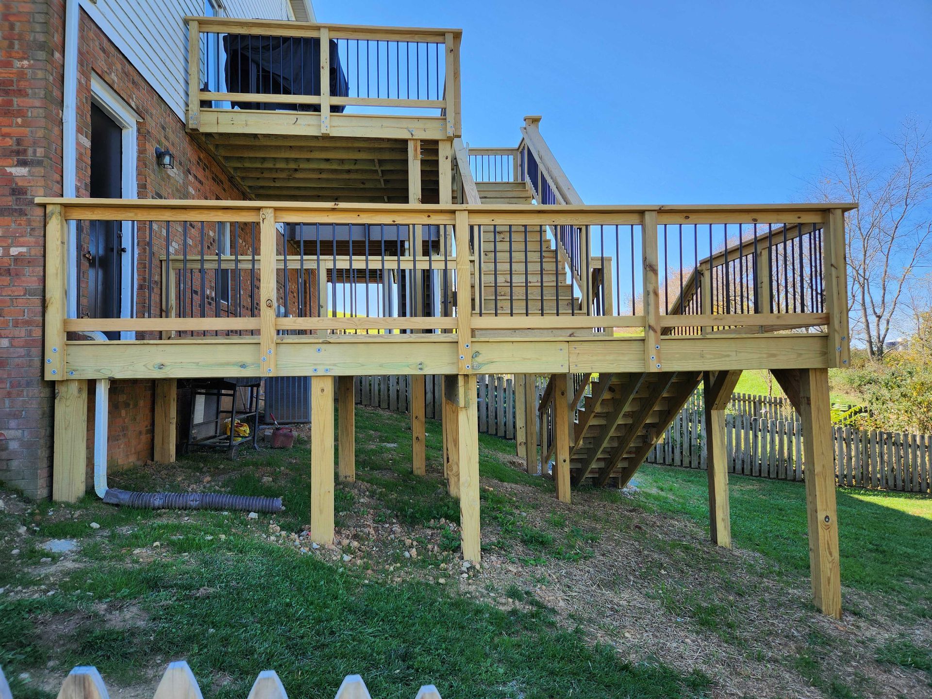 Multi-level wooden deck with black railings and stairs, attached to a brick house, on a grassy hill.