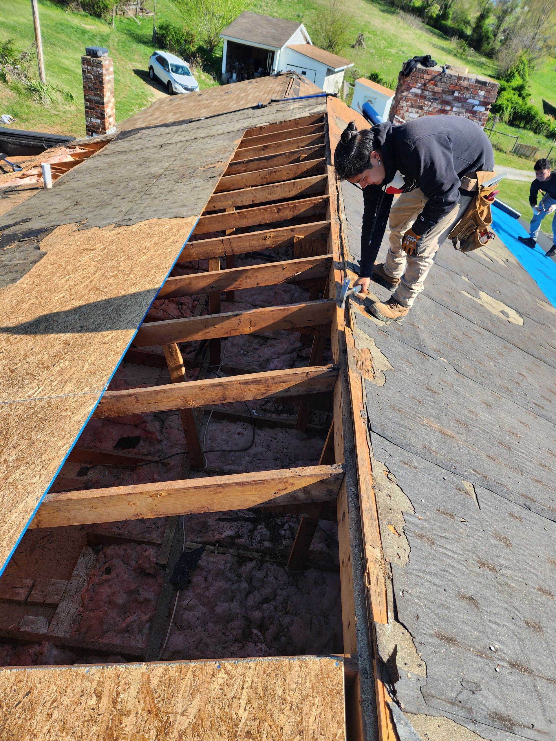 Man working on a roof, removing old shingles and exposing wooden framework. Overcast day.