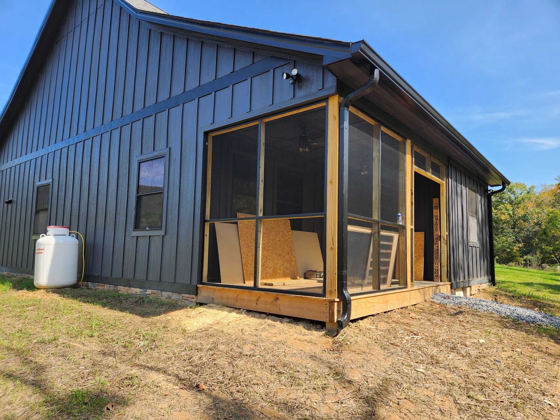 Exterior of a dark-painted building with a screened porch under construction; a propane tank sits to the left.