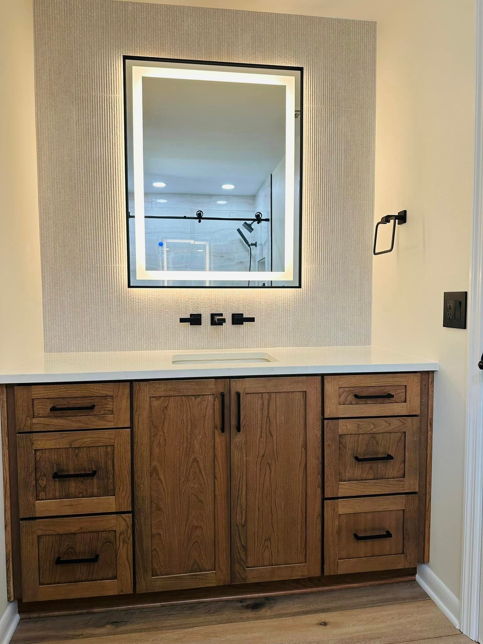 Bathroom vanity with a lighted mirror, wood cabinets, and black hardware.