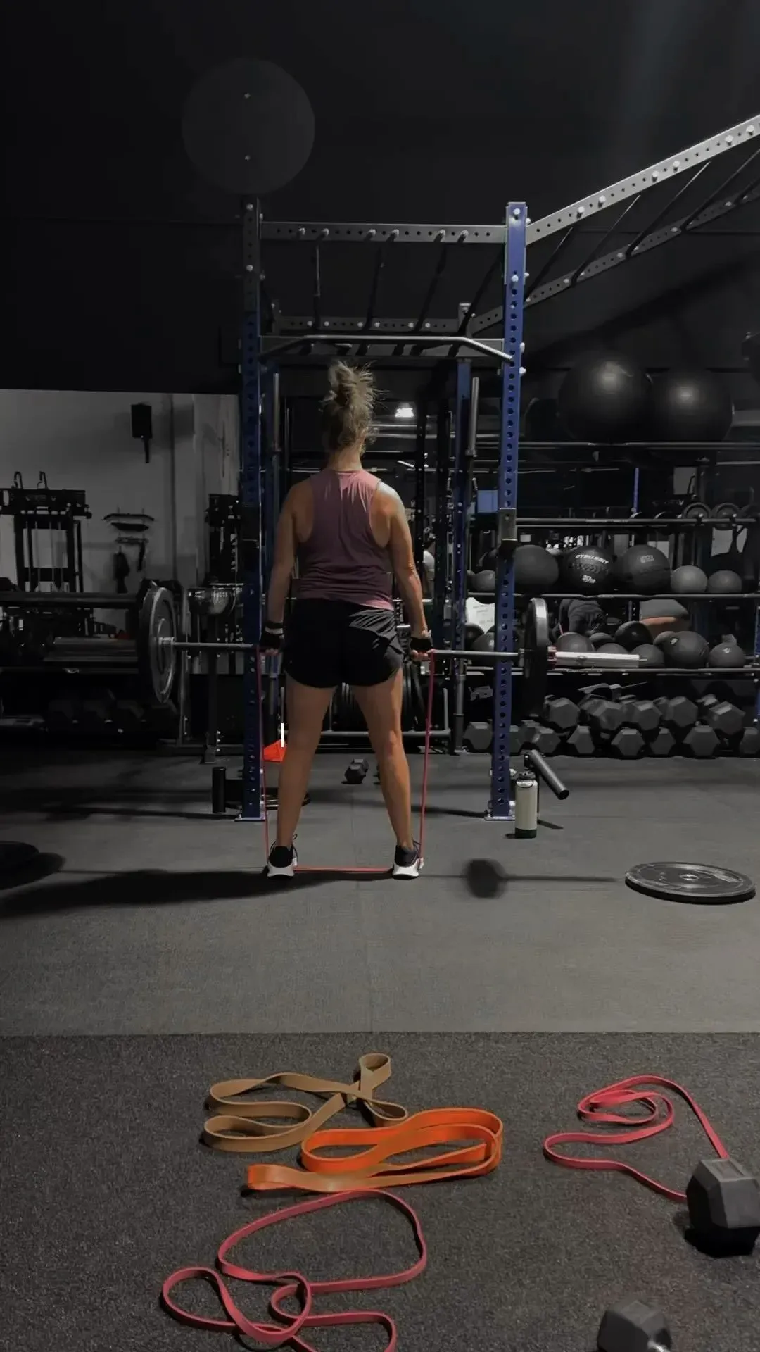 Woman in gym lifting barbell, facing away from camera. Black shorts, tank top, rack and weights in background.