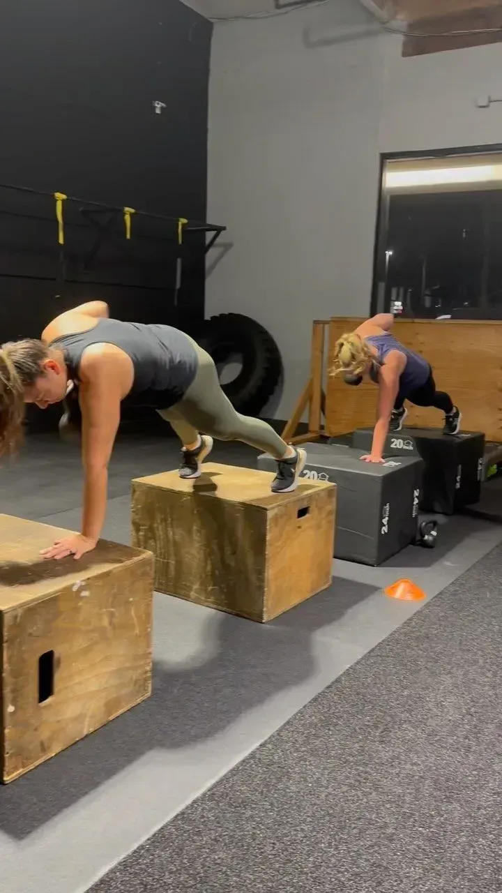 Two people exercising, doing push-ups on wooden boxes in a gym. One person's arm is raised.