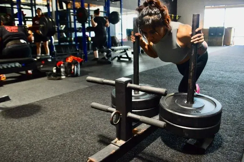 Woman pushing a weighted sled in a gym, bent over with determined expression. Black weights, rubber floor.
